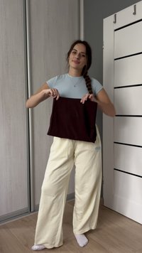 Mujer con cabello trenzado, vestida con una camiseta azul y pantalones crema, sostiene un paño oscuro doblado en una habitación con suelo de madera y puerta blanca.