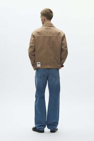 Egy férfi háttal áll, színes barna dzsekit visel, amelynek szegélyénél fehér címke található, kék farmert és fekete cipőt visel, egyszerű fehér háttér előtt.