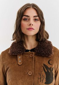 Brązowa kurtka z korduroyu z pluszowym brązowym kołnierzem, z grafiką wilka i wzorem drzew, dużymi guzikami oraz prążkowaną teksturą.