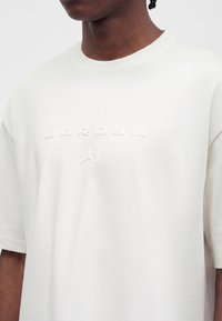 Άτομο φοράει λευκό T-shirt με στρογγυλή λαιμόκοψη και ανάγλυφο κείμενο «JORDAN» και λογότυπο Jumpman στο στήθος.