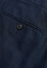 Marineblaues Gewebe mit glatter Textur, ausgestattet mit einem einzelnen schwarzen Knopf und einem gestickten Taschen Detail. Minimalistisches Design mit dezentem Glanz.