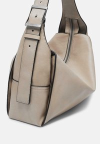 Beżowa torebka skórzana o strukturalnym kształcie, z szerokim paskiem, kontrastowym przeszyciem i zasuwanym zamknięciem po jednej stronie.