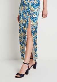 Blumiges Maxikleid in Gelb mit blauen und grünen Mustern, ausgestattet mit einem seitlichen Schlitz. Kombiniert mit schwarzen, schnürbaren, hohen Sandalen.