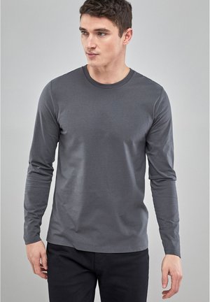 Tricou gri cu mâneci lungi, dintr-un material neted, cu decolteu rotund, design ajustat și stil minimal, asortat cu pantaloni negri.