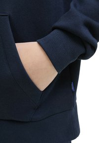 Sweatshirt bleu marine avec des poignets et un ourlet côtelés, doté d'une poche avant. Le matériau semble doux avec une texture lisse. Petite étiquette de logo visible.