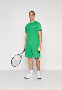 Groen sport T-shirt met witte accenten, gecombineerd met bijpassende shorts. Hij houdt een tennisracket en een bal vast, en draagt witte sneakers en sokken.