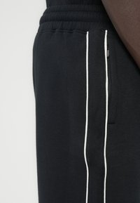 Μαύρο παντελόνι φόρμας με απαλή υφή, με λευκές ρίγες στα πλαϊνά και τσέπη με φερμουάρ κατά μήκος της αριστερής ραφής. Ορατή ελαστική μέση.