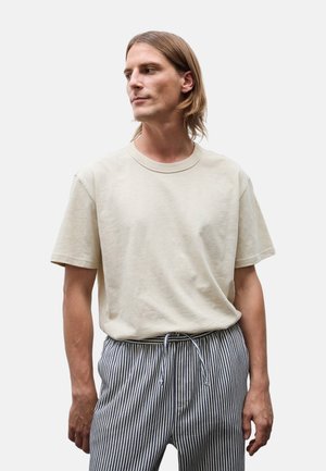 Homme aux cheveux mi-longs portant un t-shirt beige uni et un pantalon à rayures verticales noires et blanches sur un fond uni.