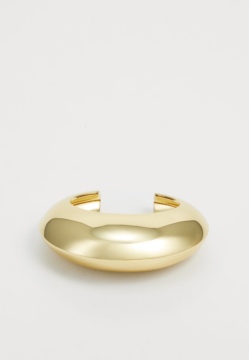 Pulsera de brazalete de metal dorado con un diseño suave y redondeado. Presenta dos aberturas en los extremos para facilitar su uso. Acabado brillante.