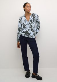 Lichtblauwe blouse met een donker abstract patroon, v-hals, lange mouwen; gecombineerd met donkerblauwe recht gesneden jeans en zwarte loafer-schoenen.
