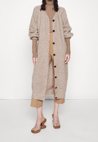 Person som bär en lång beige kavaj med svarta knappar över en brun polotröja och korta beige byxor, matchat med klacksandaler med genomskinliga remmar.