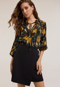 Blusa de manga larga con un patrón ikat multicolor en amarillo, verde y negro. Combinada con una falda envolvente negra ajustada con un detalle de botón.