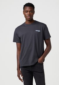 Tamsiai pilka trumpomis rankovėmis marškinėliai su apvalia apykakle ir šviesiai mėlynu „Wrangler“ logotipu kairėje krūtinės pusėje. Pagaminta iš minkštos medvilnės medžiagos.