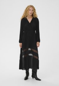 Vestido negro de manga larga con tela plisada, escote en V profundo, paneles transparentes en el dobladillo y cintura ajustada. Combinado con botas negras hasta la rodilla.