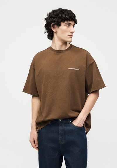 Νέος άνδρας με σγουρά σκούρα μαλλιά, φορώντας καφέ μπλουζάκι με την επιγραφή "LES BENJAMINS" και σκούρο τζιν, κοιτώντας προς τα δεξιά με το ένα χέρι στην τσέπη.