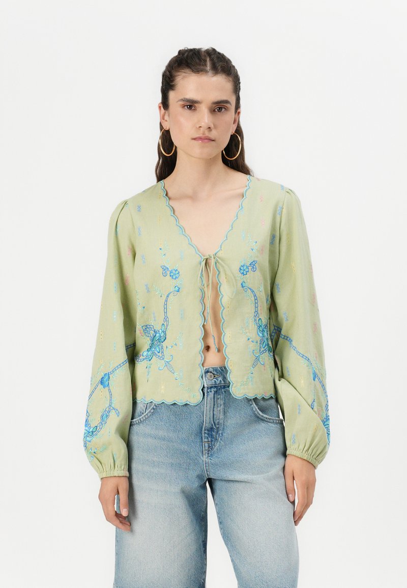 Ljusgrön blus med långa, puffiga ärmar, vågiga kanter och blå blommönster broderade i mönster. Knut vid ringningen, tillsammans med jeans.
