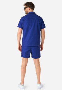 Camicia blu navy a maniche corte abbinata a pantaloni corti coordinati. Tessuto morbido, design semplice e orlo arrotolato. Sneakers bianche completano il look.