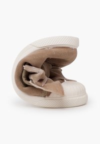 Flexibler beiger Sneaker mit weißer Gummisohle und Zehenkappe, auf die Hälfte gefaltet, um Weichheit und Biegefähigkeit zu demonstrieren.