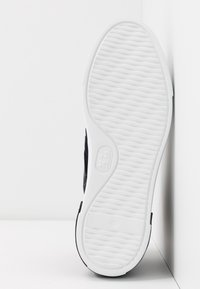 Mustavalkoinen urheilukengän pohja, jossa on teksturoitu valkoinen kumipohja, jossa on aaltoilevia uurteita pidon varmistamiseksi ja pyöreä logo-tyyppinen yksityiskohta lähellä kantapäätä.