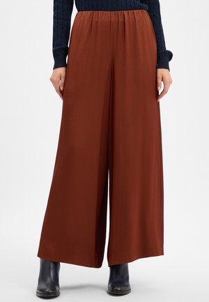 Vrouw die wijde roestkleurige broek, een marineblauwe trui en zwarte enkellaarzen draagt, staande tegen een effen achtergrond.