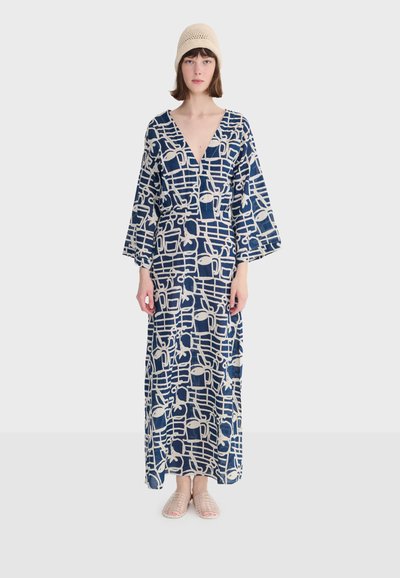 Kobieta ubrana w długą granatowo-beżową sukienkę w wzory z szerokimi rękawami, beżową dzianinową czapkę i beżowe plecione buty wsuwane, stojąca na tle białego tła.