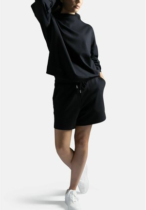 Henkilö seisoo ristissä jaloin, hänellä on päällään musta pitkähihainen paita, mustat shortsit ja valkoiset tennarit, toinen käsi taskussa, valkoista taustaa vasten.