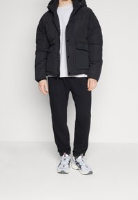 Man som bär en svart puffjacka, ljusgrå skjorta, svarta joggingbyxor och vita sneakers, står mot en enkel ljus bakgrund.