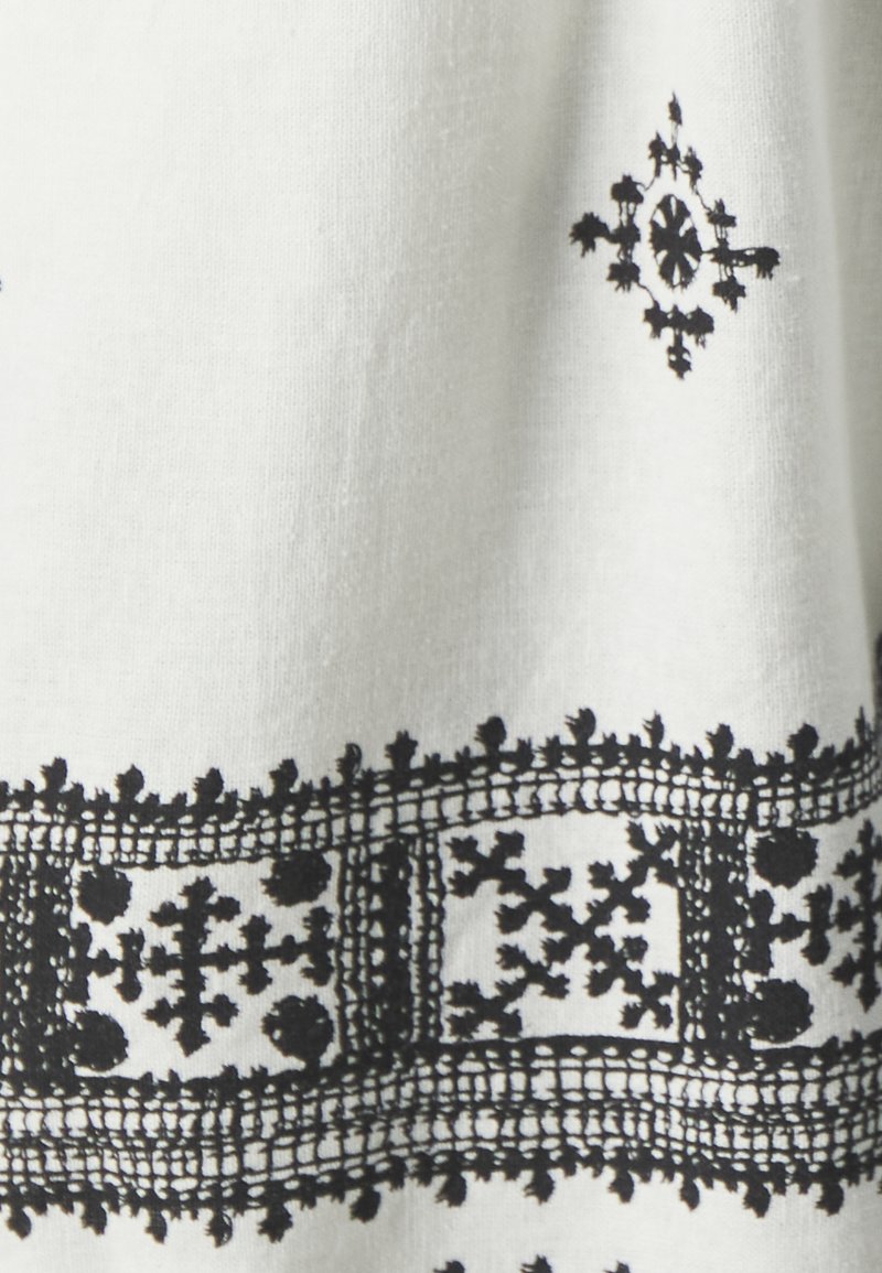 Textil blanco con bordados negros intrincados que presentan patrones geométricos y bordes decorativos. Textura suave con colores contrastantes.