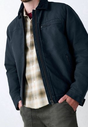 Mann trägt eine aufgeknöpfte schwarze Jacke über einem beige karierten Hemd, mit Händen in den Taschen, zeigt ein lässiges, geschichtetes Outfit.