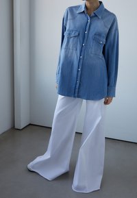 Camisa de mezclilla azul claro con dos bolsillos en el pecho, cierre de botones, mangas largas, combinada con pantalones anchos blancos. Textura suave, diseño informal.