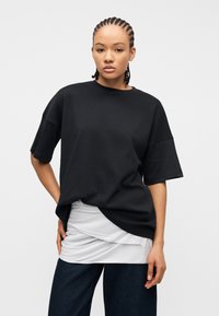 Νεαρός με πλεγμένα μαλλιά, φορώντας ένα χαλαρό μαύρο T-shirt πάνω από λευκό στρωμένο τοπ και σκούρο μπλε παντελόνι, στέκεται μπροστά σε ένα απλό φόντο.