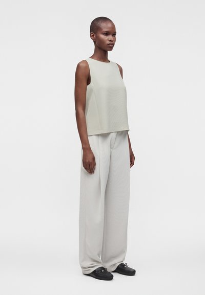 Personne debout de profil portant un haut beige ample et sans manches, un pantalon clair à jambes larges et des chaussures noires sur un fond uni.