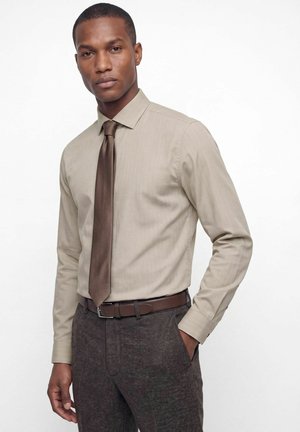 Homme portant une chemise beige, une cravate marron, une ceinture marron et un pantalon marron foncé, debout avec une main dans la poche sur fond blanc.