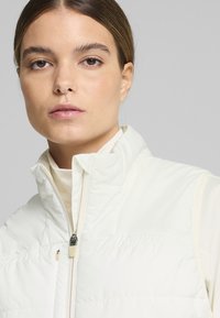 White puffer vest with a high collar and zipper closure, featuring a quilted design and smooth texture, worn over a light-colored top.