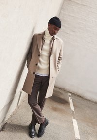 Beige wollen overcoat met zwarte knopen over een crèmekleurige coltrui. Donkerbruine gestreepte broek en zwarte stevige schoenen. Zwarte gebreide beanie.