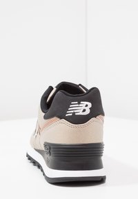 Tenisky New Balance v béžové a černé barvě. Mají kožený a síťovinový materiál, kulatý tvar, polstrovaný límec a výrazné logo na patě.