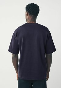 Man med kort lockigt hår som bär en enkel mörk lila t-shirt med korta ärmar, stående vänd bort mot en ljus bakgrund.