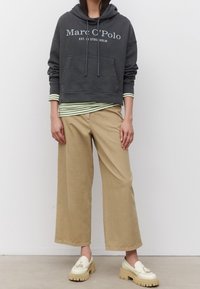 Grijze cropped capuchontrui met voorzak en logo over een groen gestreept shirt met lange mouwen, gecombineerd met beige broek met wijde pijpen en crèmekleurige loafers.