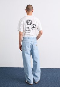 Hvid t-shirt med sorte grafiske designs på ryggen, parret med lyseblå brede jeans. Blødt materiale og afslappet pasform.