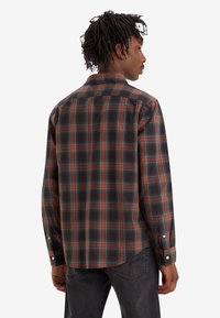 Hombre de espaldas a la cámara que lleva una camisa de cuadros marrón, naranja y azul de manga larga y vaqueros gris oscuro frente a un fondo blanco.