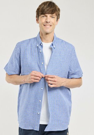 Homme aux cheveux châtain clair souriant en boutonnant une chemise à manches courtes à carreaux bleu et blanc par-dessus un t-shirt blanc sur un fond uni.