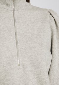 Suéter gris claro con cuello alto y cierre de cremallera. Presenta un tejido texturizado y acentuaciones de hombros abullonados. Diseño suave y casual.