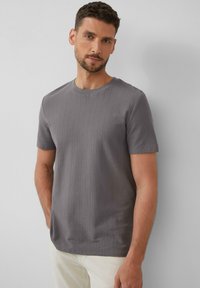 Graues, geripptes Kurzarm-T-Shirt mit Rundhalsausschnitt. Der Stoff wirkt weich und hat eine dezente Textur. Getragen mit hellen Hosen.