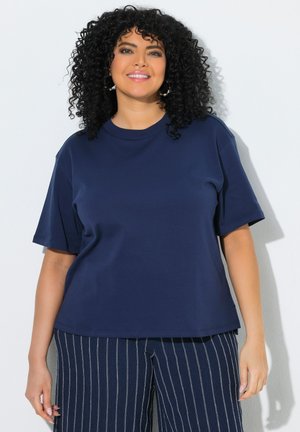 Smilende kvinde med krøllet sort hår iført en ensfarvet marineblå T-shirt og marineblå bukser med hvide lodrette striber, stående mod en hvid væg.