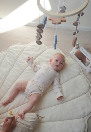 Niemowlę leży na kremowej, pikowanej macie pod drewnianym karuzelą z wiszącymi kształtami, patrzy w stronę osoby trzymającej smoczek w pobliżu.