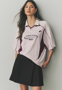 Un t-shirt oversized rose et bourgogne avec "MILAN" et "ITALIE" imprimés, associé à une jupe plissée noire. Manches courtes, décolleté en V.