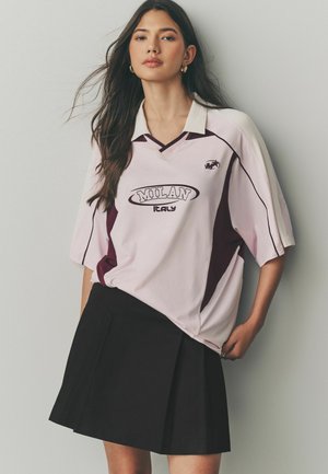 Un t-shirt oversized rose et bourgogne avec "MILAN" et "ITALIE" imprimés, associé à une jupe plissée noire. Manches courtes, décolleté en V.