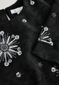 Robe en tissu noir avec des motifs floraux en éclat d'étoile composés de sequins argentés et de perles sur le corsage et la manche, présentant un gros plan sur la texture et les détails.