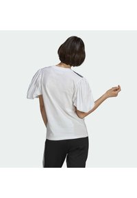 Camiseta blanca con mangas abullonadas, con un escote redondo y un pequeño detalle de raya negra en los hombros. Lleva puesta con pantalones negros.