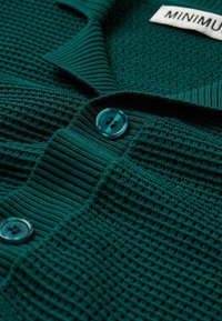 Cardigan en tricot turquoise avec un motif texturé, un col côtelé et une fermeture à boutons ronds et brillants. L'étiquette indique "MINIMUM."